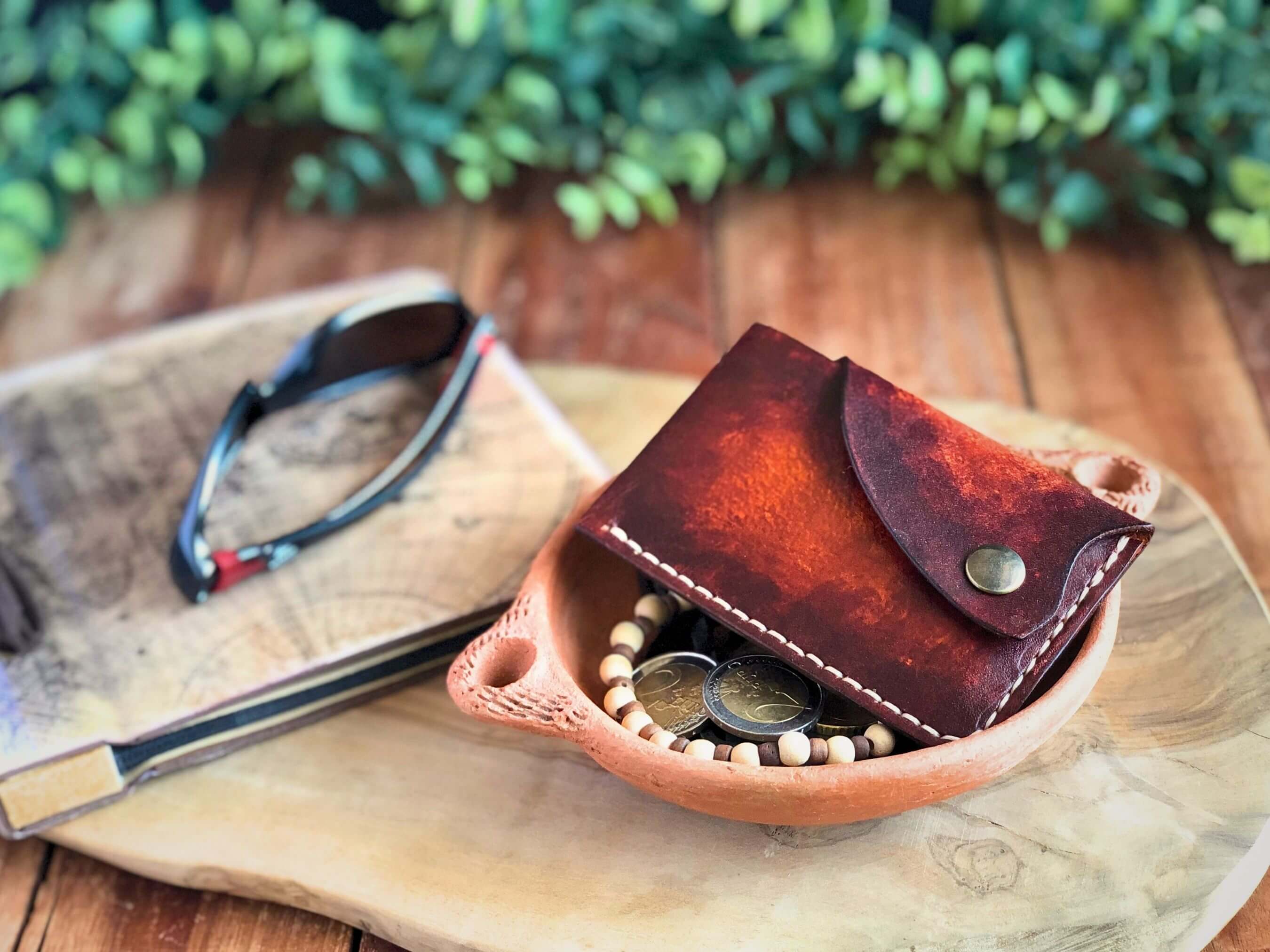 Brown leather wallet with snap closure laying with with coins on a wooden surface with sunglasses and a plant in the background