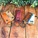Three leather emergency keychain wallets on a wooden surface with greenery above.