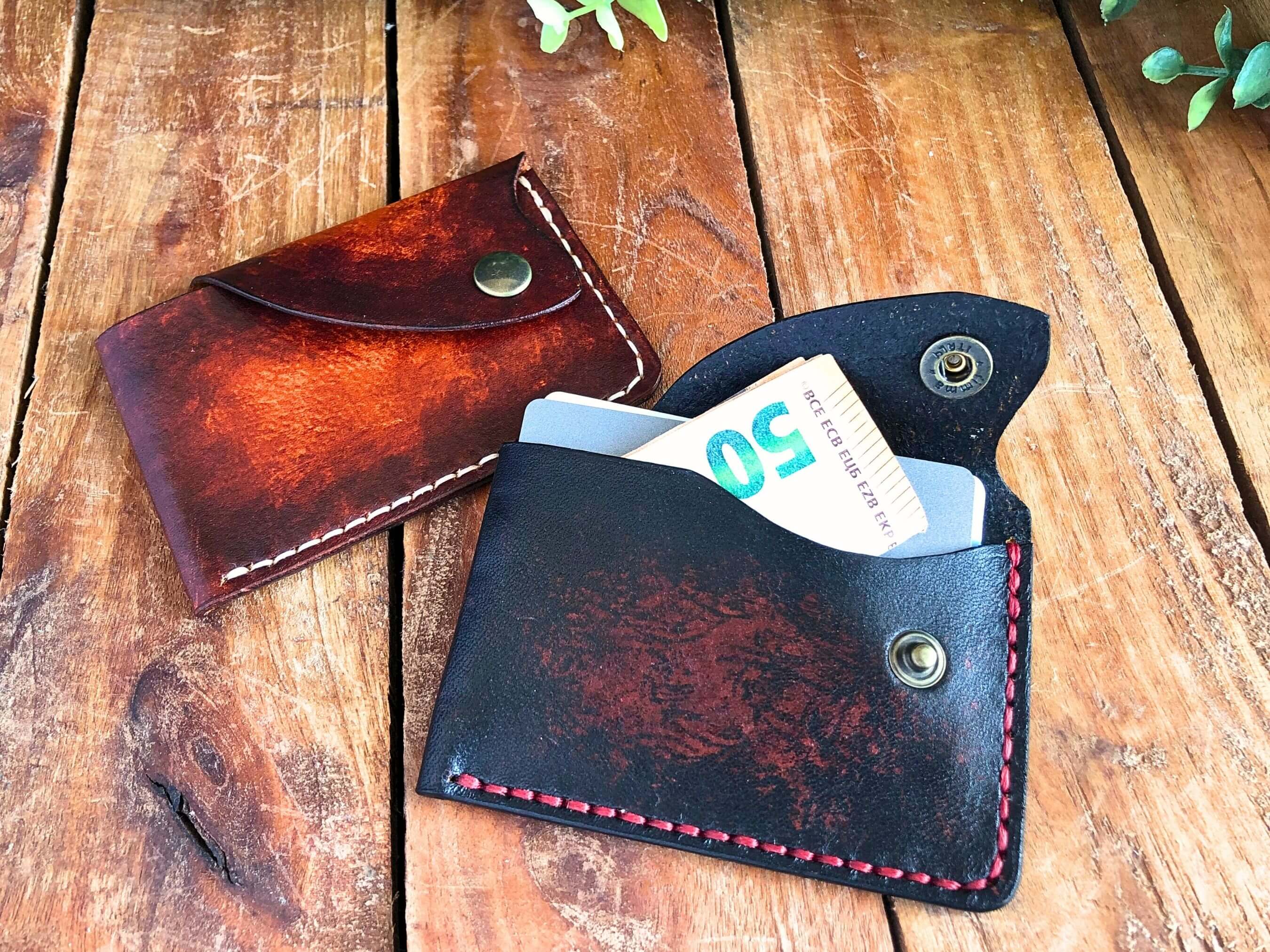 Two slim leather card wallets with snap closure on a wooden surface with one containing a 50 euro bill.