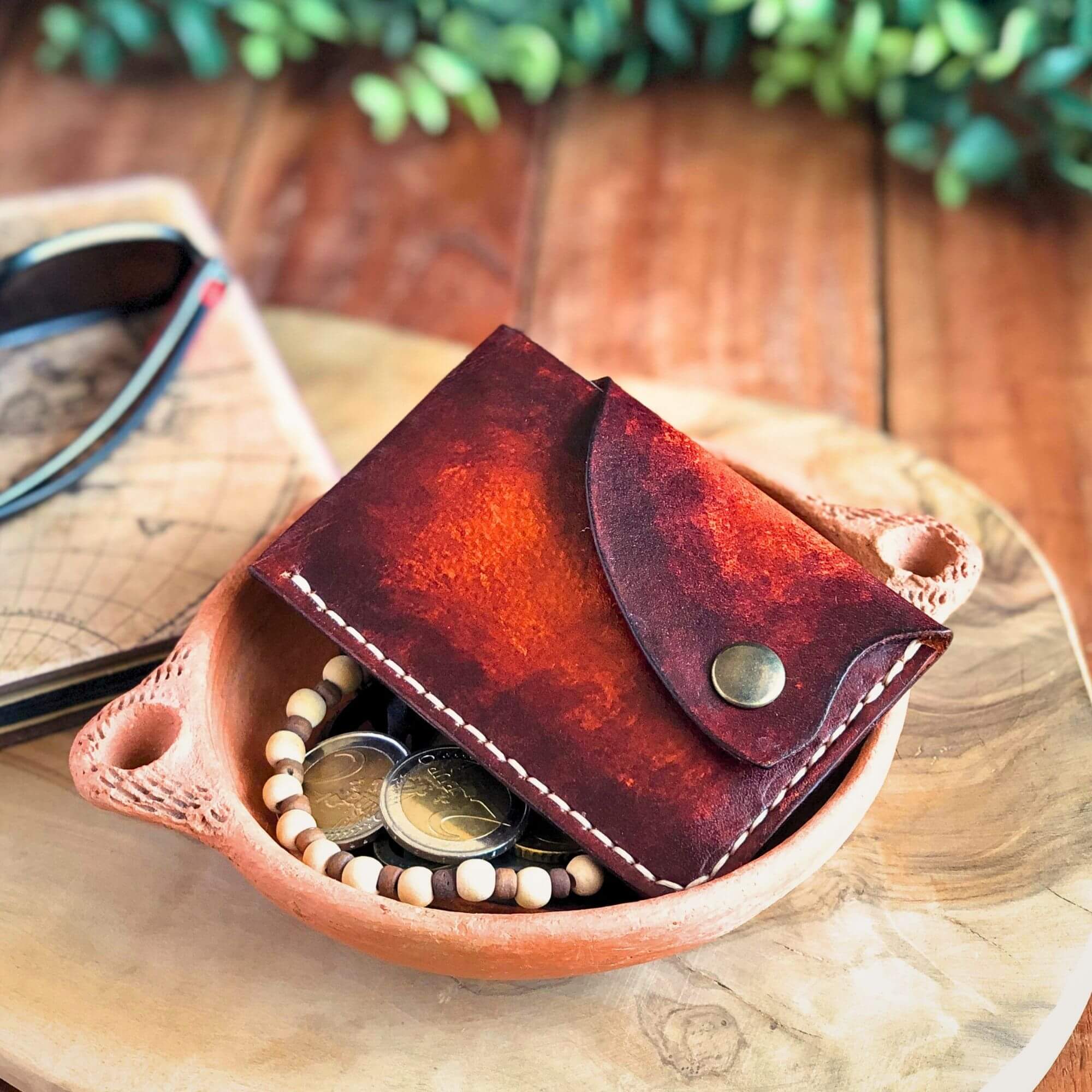Brown leather wallet with snap closure laying with with coins on a wooden surface with sunglasses and a plant in the background
