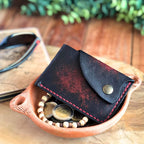 Black leather card wallet with snap closure laying with with coins on a wooden surface with sunglasses and a plant in the background