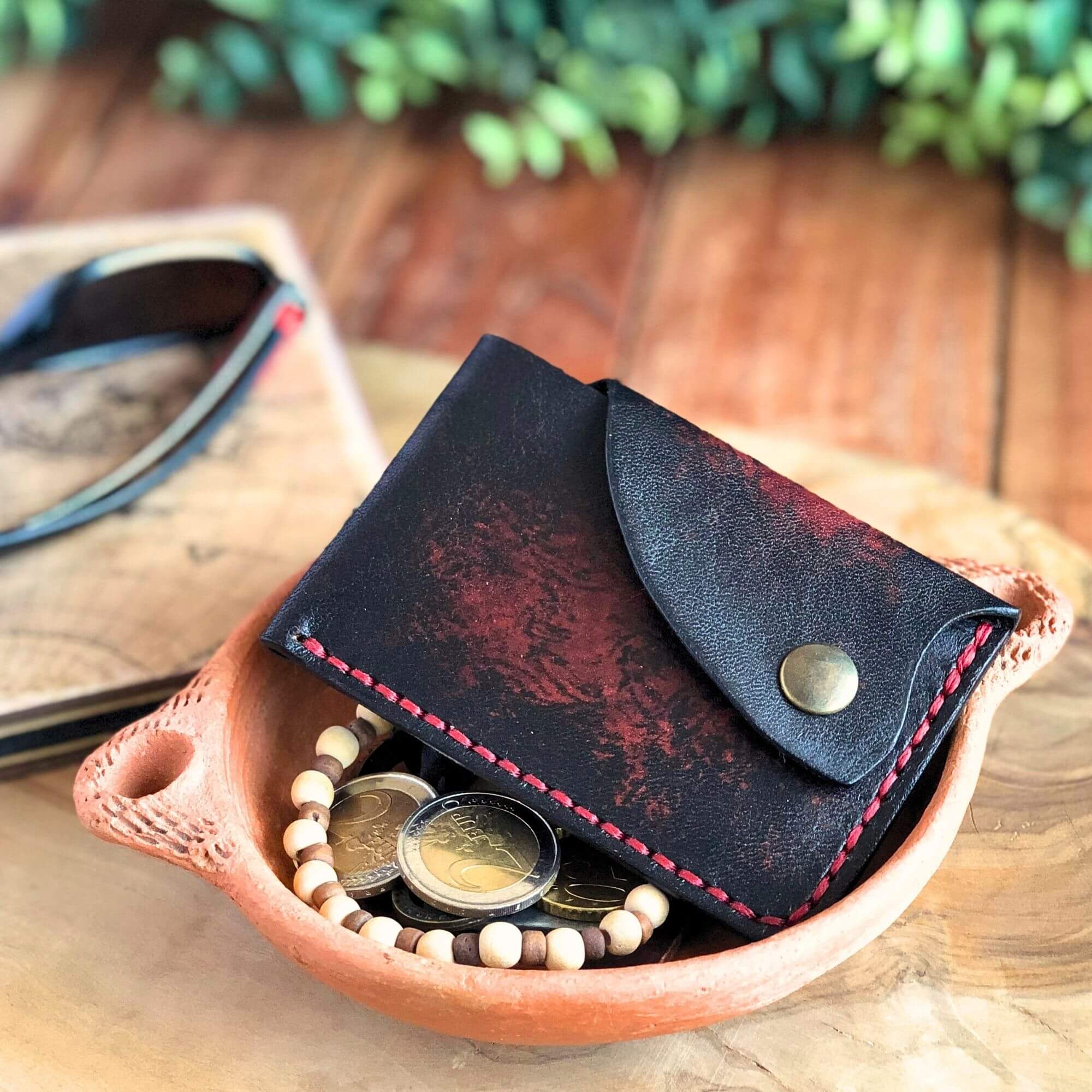Black leather card wallet with snap closure laying with with coins on a wooden surface with sunglasses and a plant in the background