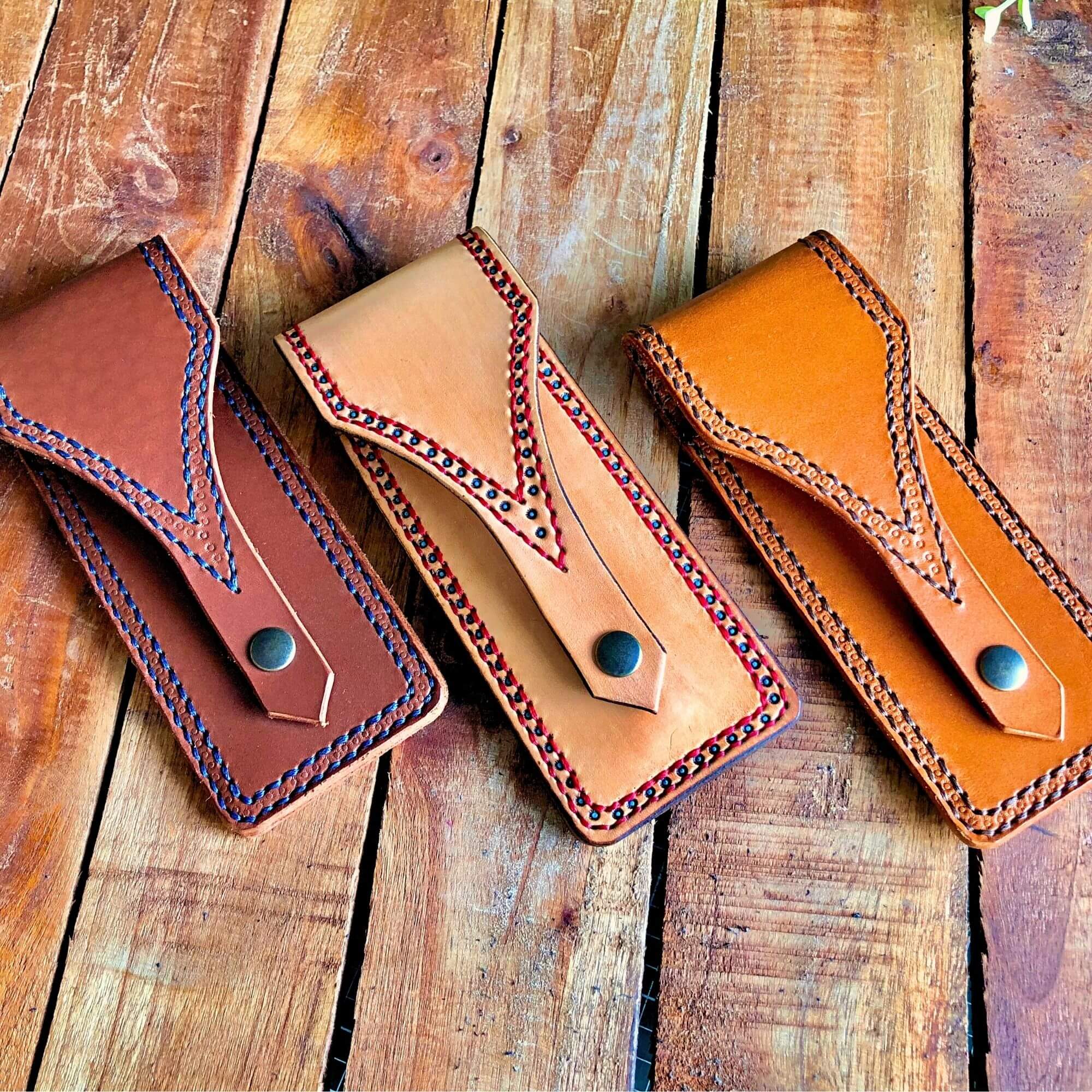 Three leather pen pouches in brown, tan, and natural with decorative stitching on a wooden surface