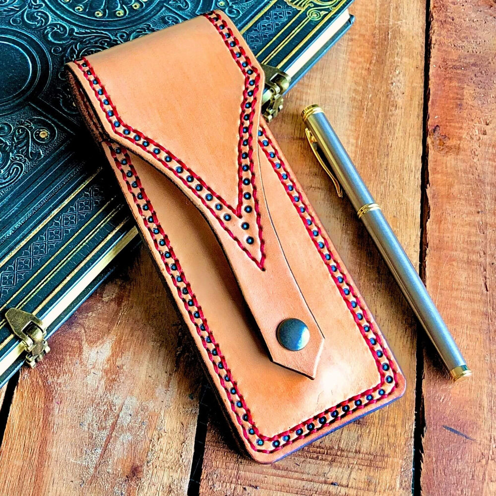 Leather pen pouch with decorative stitching on a wooden surface with a pen and notebook.