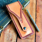 Leather pen pouch with decorative stitching on a wooden surface with a pen and notebook.