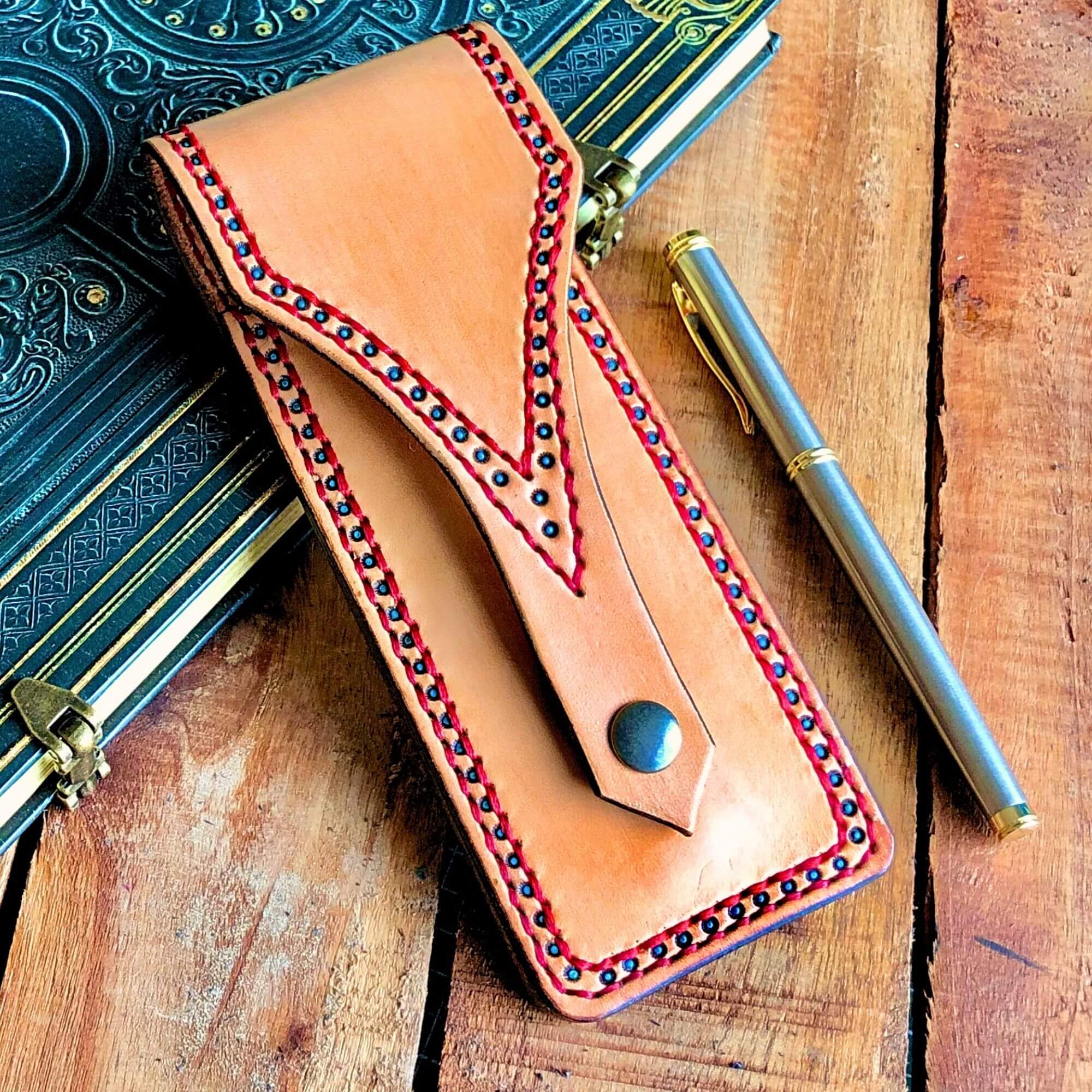 Leather pen pouch with decorative stitching on a wooden surface with a pen and notebook.