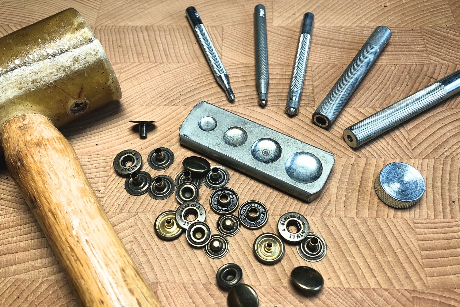 snaps laid out on a wooden surface together with the tools required to install them in leather, such as a rawhide mallet and various setter tools
