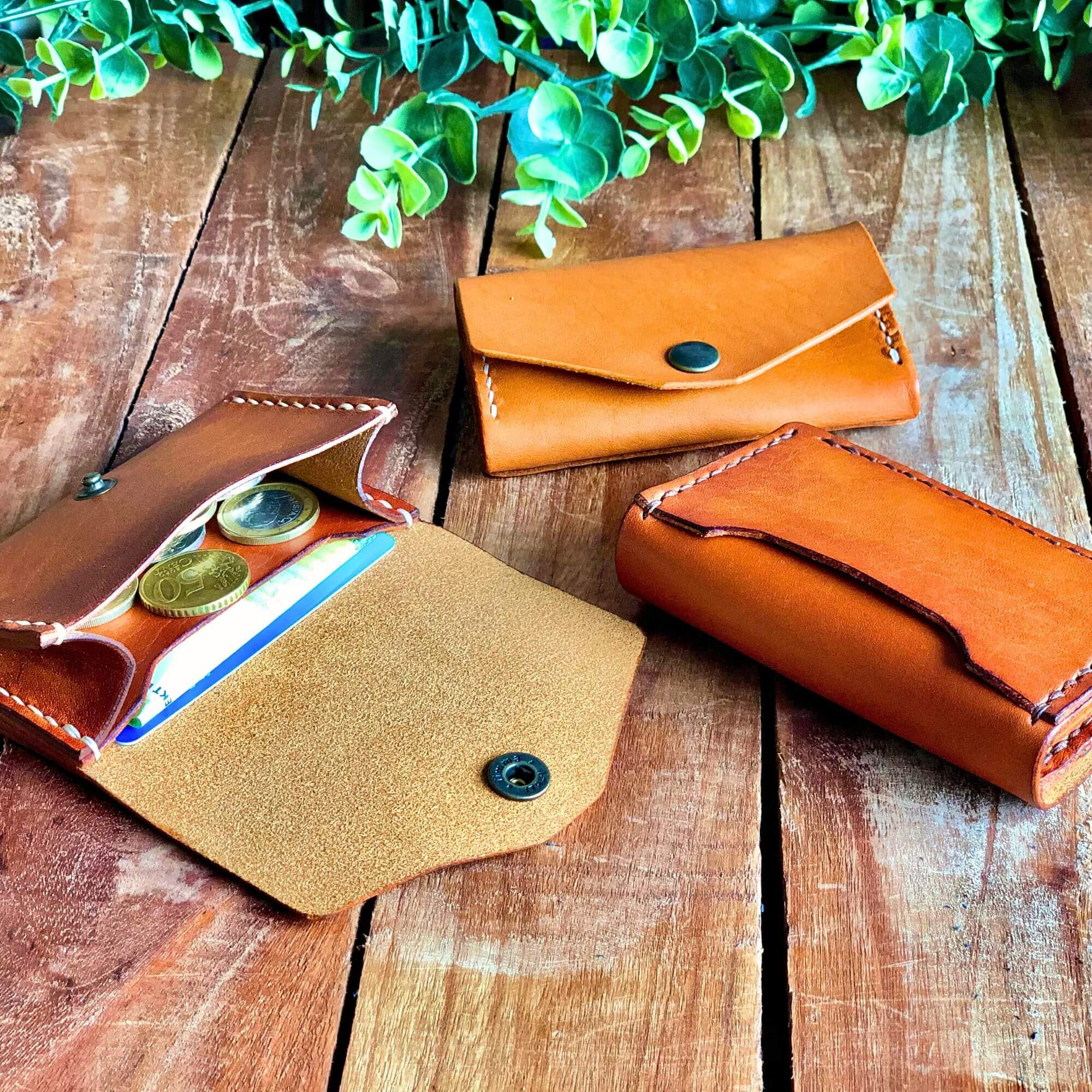 Three tan leather wallets on a wooden surface with green leaves in the background